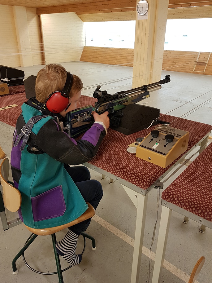 Otto is sitting on a chair with an air rifle in his hand pointing towards the board